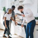 Professional bearded chubby man in formal outfit with clipboard inspecting oven together with attentive ethnic couple while standing at modern sunny apartment